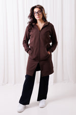 Modelka w długiej brązowej bluzie Long BalticBird z polskiej bawełny, zapinanej na suwak, z kieszeniami i wydłużonym tyłem; stylizacja z czarnymi spodniami loose i białymi sneakersami; sesja w Gdańsku; odzież sportowa dla kobiet 30+ dopasowana do sylwetek jabłko, gruszka i kolumna.