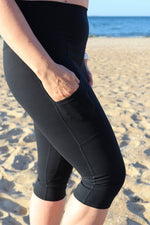 Modelka na plaży w czarnym topie sportowym i czarnych legginsach typu 3/4  za kolano stoi boso na piasku, w tle morze i jasne niebo.