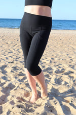 Modelka na plaży w czarnym topie sportowym i czarnych legginsach typu 3/4  za kolano stoi boso na piasku, w tle morze i jasne niebo.